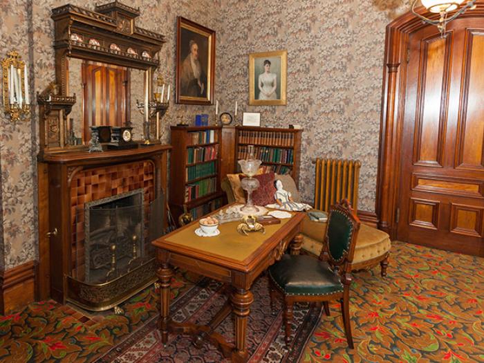 Alexander Ramsey House livingroom. A dark maple fireplace with tile with a table with oil lamp and leather chair. Bookshelves and very busy wallpaper